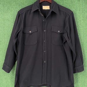 Könnte beinhalten: Ein schwarzes Langarmhemd mit zwei Brusttaschen und Knopfverschlüssen. Das Hemd hat einen Kragen und hängt an einem Holzbügel. Der Stoff scheint ein mittelschweres Material zu sein.