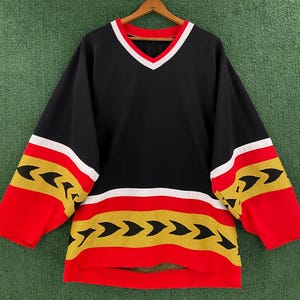 May include: Black hockey jersey with red and gold trim. The jersey has a V-neck and a white stripe across the chest. The sleeves and bottom of the jersey have a pattern of gold arrows on a red background.