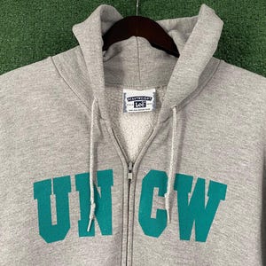 May include: A grey zip-up hoodie featuring the teal letters "UNCW" across the chest. The hoodie has a drawstring hood and a "Heavyweight Lee" label. The zipper is silver.