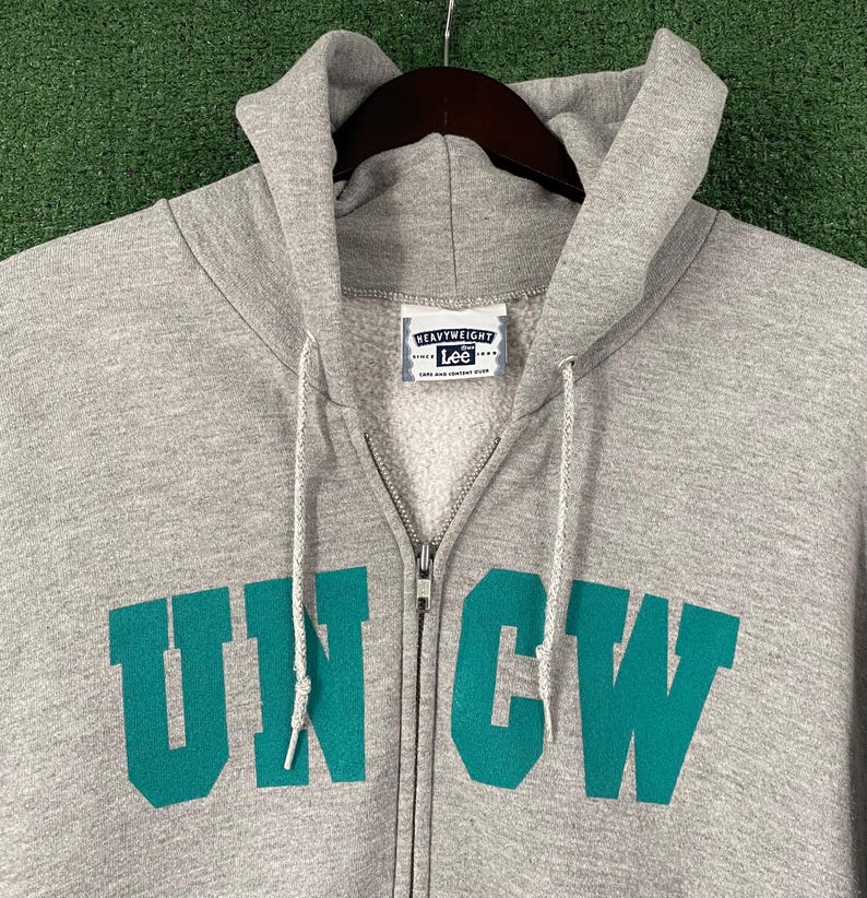 May include: A grey zip-up hoodie featuring the teal letters "UNCW" across the chest. The hoodie has a drawstring hood and a "Heavyweight Lee" label. The zipper is silver.