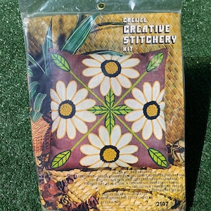 May include: A sealed package containing a Crewel Creative Stitchery Kit. The kit features a pillow design with four large white daisies with yellow centers and green leaves on a maroon background. The finished pillow size is 13 inches by 13 inches.