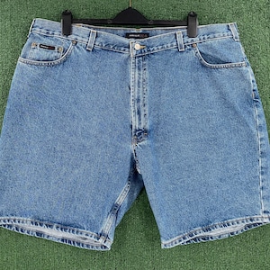 May include: Light blue denim shorts with a classic five-pocket design. The shorts have a button and zipper closure, belt loops, and a small pocket on the front. The shorts are hanging on a black hanger against a green background.