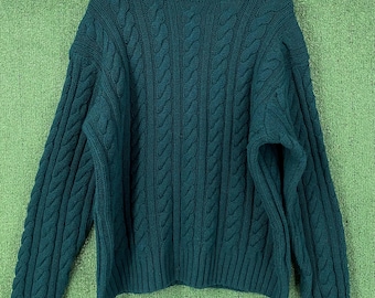 May include: A teal cable-knit turtleneck sweater. The sweater features a ribbed turtleneck, cuffs, and hem. The cable-knit design covers the front, back, and sleeves. The sweater is hanging on a wooden hanger against a green background.