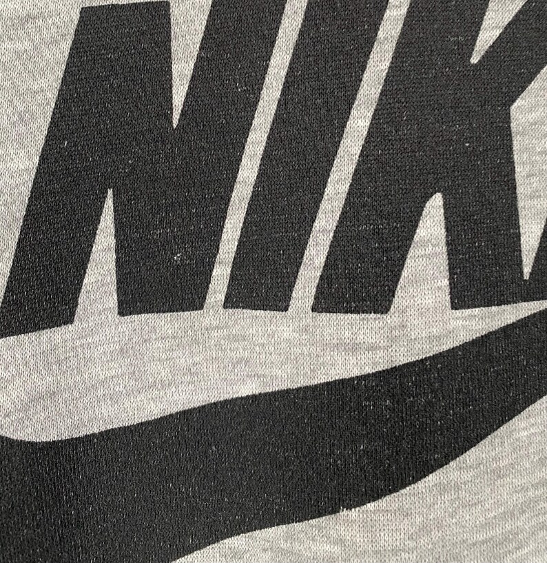 May include: Close-up of a gray fabric with the black Nike logo. The logo is large and centered, with the word "NIKE" prominently displayed. The fabric has a slightly textured appearance.
