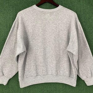 May include: A heather gray sweatshirt with a crew neck and long sleeves. The sweatshirt is made of a soft material and has ribbed cuffs and hem. The garment is displayed on a wooden hanger against a green background.
