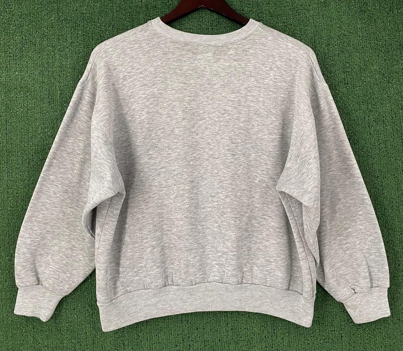 May include: A heather gray sweatshirt with a crew neck and long sleeves. The sweatshirt is made of a soft material and has ribbed cuffs and hem. The garment is displayed on a wooden hanger against a green background.