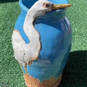 Puede incluir: Un jarrón de cerámica con un esmalte azul vibrante y un diseño de garza blanca. La cabeza y el cuello de la garza se extienden sobre el borde del jarrón. La base del jarrón tiene un acabado marrón texturizado. Una pieza decorativa única.
