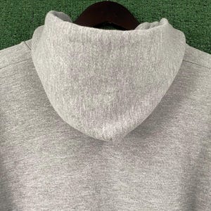 May include: Close-up of a grey hoodie with a high collar. The hood is made of a soft, textured fabric. The hoodie is hanging on a dark brown wooden hanger against a green background.