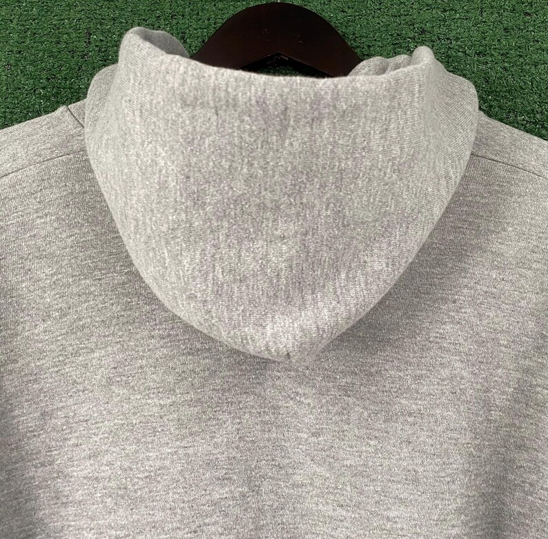 May include: Close-up of a grey hoodie with a high collar. The hood is made of a soft, textured fabric. The hoodie is hanging on a dark brown wooden hanger against a green background.