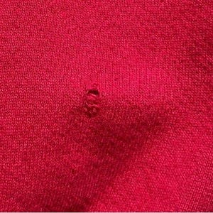 Może przedstawiać: Zbliżenie na jasnoczerwony dzianinowy materiał z małą dziurą. Tkanina ma teksturowany wygląd z widocznymi ściegami dziewiarskimi. Otw&oacute;r znajduje się w pobliżu środka obrazu, odsłaniając strukturę materiału.