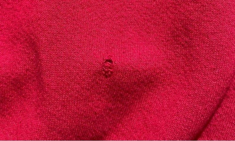 Może przedstawiać: Zbliżenie na jasnoczerwony dzianinowy materiał z małą dziurą. Tkanina ma teksturowany wygląd z widocznymi ściegami dziewiarskimi. Otw&oacute;r znajduje się w pobliżu środka obrazu, odsłaniając strukturę materiału.