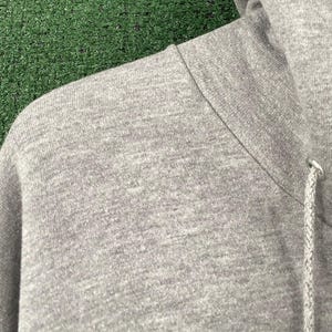 May include: Close-up of a grey hoodie, showing the shoulder and hood. The fabric has a textured appearance. The drawstring and metal eyelet are visible. The background is green.