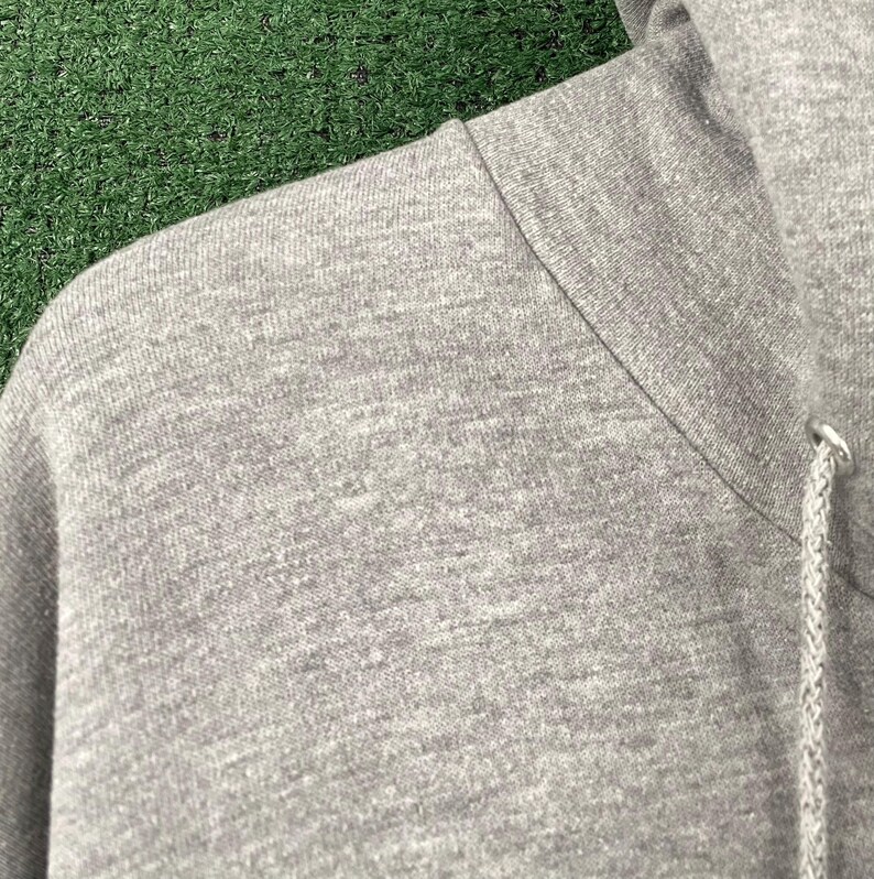 May include: Close-up of a grey hoodie, showing the shoulder and hood. The fabric has a textured appearance. The drawstring and metal eyelet are visible. The background is green.