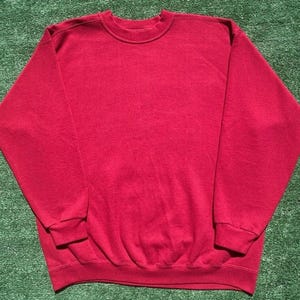 Może przedstawiać: Gładka czerwona bluza z okrągłym dekoltem. Top z długim rękawem wykonany jest z miękkiego materiału i ma prążkowany d&oacute;ł i mankiety. Bluza leży płasko na zielonej powierzchni.