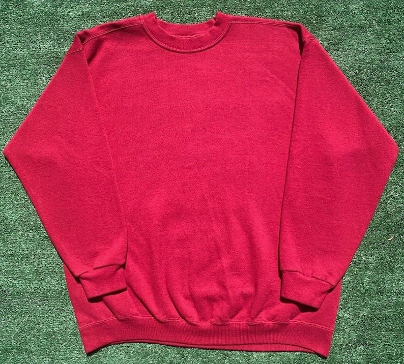 Może przedstawiać: Gładka czerwona bluza z okrągłym dekoltem. Top z długim rękawem wykonany jest z miękkiego materiału i ma prążkowany d&oacute;ł i mankiety. Bluza leży płasko na zielonej powierzchni.