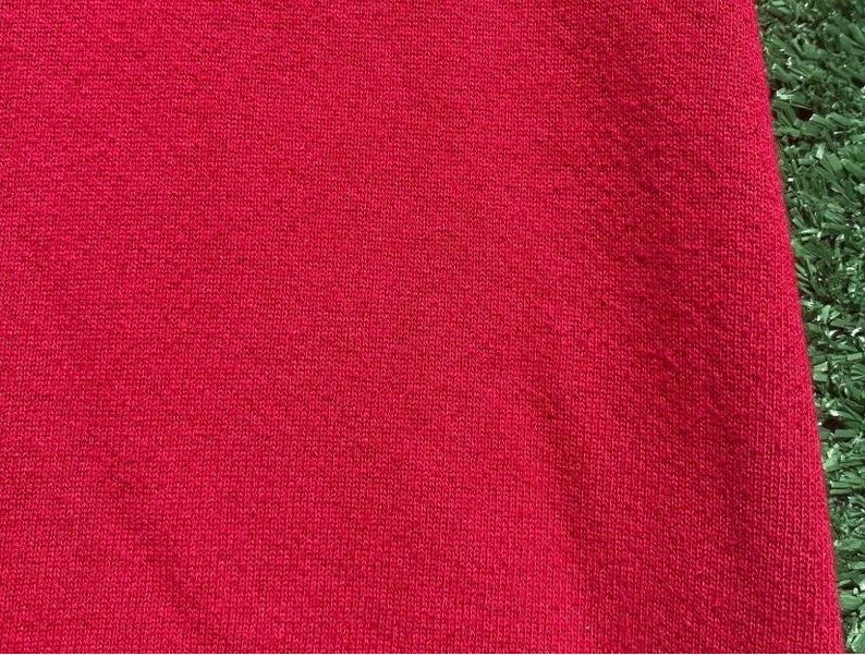 Może przedstawiać: Zbliżenie fragmentu czerwonej tkaniny. Materiał ma teksturowany wygląd z widocznym wzorem dzianiny. Kolor jest żywy, jednolity czerwony, wypełniający kadr. Tkanina wydaje się miękka i prawdopodobnie dzianinowa.