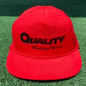 Pode incluir: Boné de beisebol vermelho brilhante com o texto "QUALITY Racing Team" em preto. O boné tem uma aba curva e um detalhe de corda trançada na base. Feito de um material leve, possivelmente nylon.