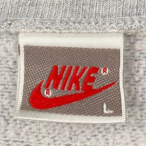 May include: Close-up of a gray sweatshirt tag with the red Nike logo and the letter "L". The tag is rectangular with a white border and is sewn onto the gray fabric of the sweatshirt.