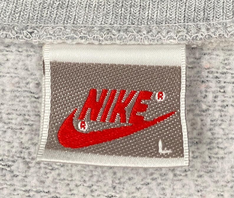 May include: Close-up of a gray sweatshirt tag with the red Nike logo and the letter "L". The tag is rectangular with a white border and is sewn onto the gray fabric of the sweatshirt.