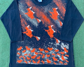 Peut inclure: Un t-shirt noir à manches longues avec un imprimé Halloween orange et blanc. L'imprimé représente des sorcières volant sur des balais et des chauves-souris.