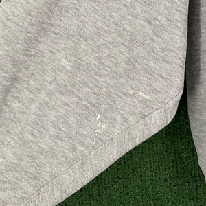 May include: Close-up of a gray sweatshirt sleeve. The fabric has a textured appearance with a few small white paint marks. The sleeve is cuffed at the wrist. The background is a green surface.