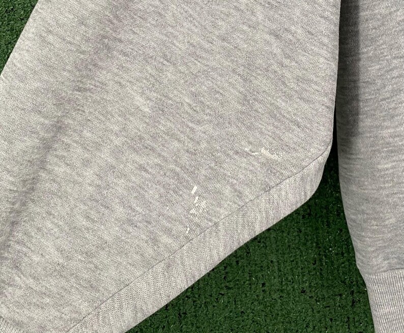 May include: Close-up of a gray sweatshirt sleeve. The fabric has a textured appearance with a few small white paint marks. The sleeve is cuffed at the wrist. The background is a green surface.