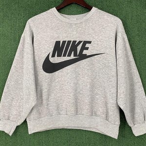 May include: A light gray sweatshirt with the black Nike logo across the chest. The logo includes the brand name and the iconic swoosh. The sweatshirt has a crew neck and long sleeves.