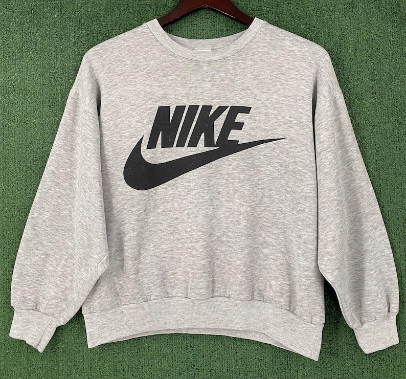 May include: A light gray sweatshirt with the black Nike logo across the chest. The logo includes the brand name and the iconic swoosh. The sweatshirt has a crew neck and long sleeves.