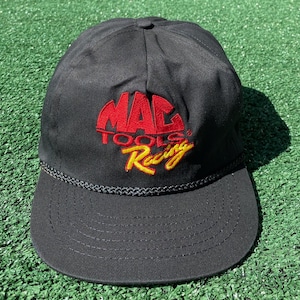 Könnte beinhalten: Schwarze Baseballkappe mit dem gestickten Text "MAC TOOLS Racing" in Rot und Gelb. Die Kappe hat einen gebogenen Schirm und eine geflochtene Kordel am Rand des Schirms.