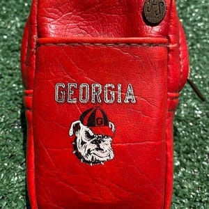 May include: A red textured leather pouch featuring the word GEORGIA printed in white above a graphic of a bulldog wearing a red and black cap. A circular metal emblem with the letters JFS is attached to the upper right corner of the pouch.