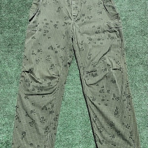 Könnte beinhalten: Grün-braune Camouflage-Cargo-Hose mit Kordelzugbund und mehreren Taschen. Die Hose hat ein sich wiederholendes quadratisches Muster mit dunkleren grünen Formen. Die Hose liegt auf einer grünen Oberfläche.