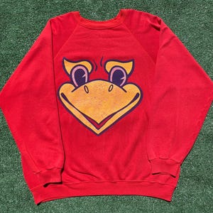 May include: A bright red long-sleeve sweatshirt featuring a large, centered graphic of a stylized yellow bird face with dark purple outlines, large eyes, and a prominent beak. The garment has a crew neckline, ribbed cuffs, and a ribbed hem.