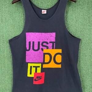 Puede incluir: Camiseta sin mangas negra de Nike con la frase "JUST DO IT" en un diseño colorido y en bloques. El texto es morado, naranja, amarillo y negro. Un pequeño logotipo rojo de Nike en la parte superior.