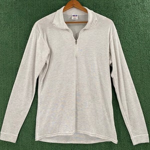 Puede incluir: Una camisa gris claro de manga larga con cuello con cremallera de un cuarto. La camisa está hecha de un material suave y texturizado y tiene dobladillo y puños acanalados. La camisa está colgada en una percha de madera sobre un fondo verde.