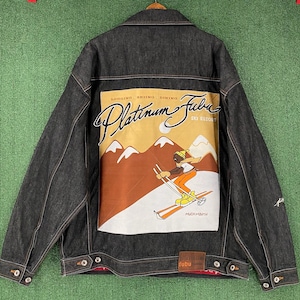 May include: Dark denim jacket with a colourful graphic on the back featuring a skier, mountains, and the words "Platinum Fubu Ski Resort." The jacket has button closures and the Fubu logo.
