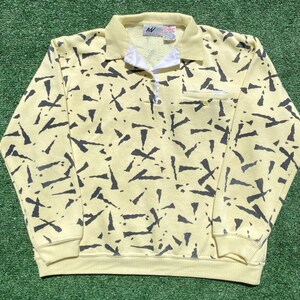 May include: A pale yellow long-sleeve pullover shirt with a dark gray abstract jagged pattern. The shirt features a ribbed collar, a white button-up placket, and a small chest pocket with a single button. A brand label is visible at the neck.