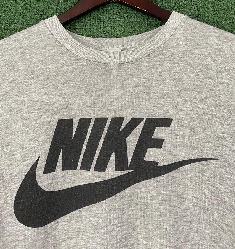 May include: A heather gray crew neck sweatshirt with the black Nike logo and swoosh. The sweatshirt is hanging on a wooden hanger against a green background.