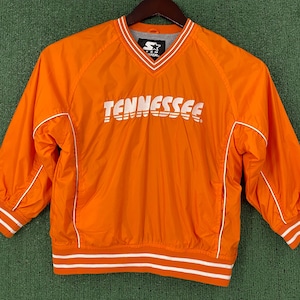 May include: Orange pullover windbreaker with white accents and the word "TENNESSEE" across the chest. Features a V-neck, Starter logo, and striped cuffs and hem. A sporty, lightweight jacket.