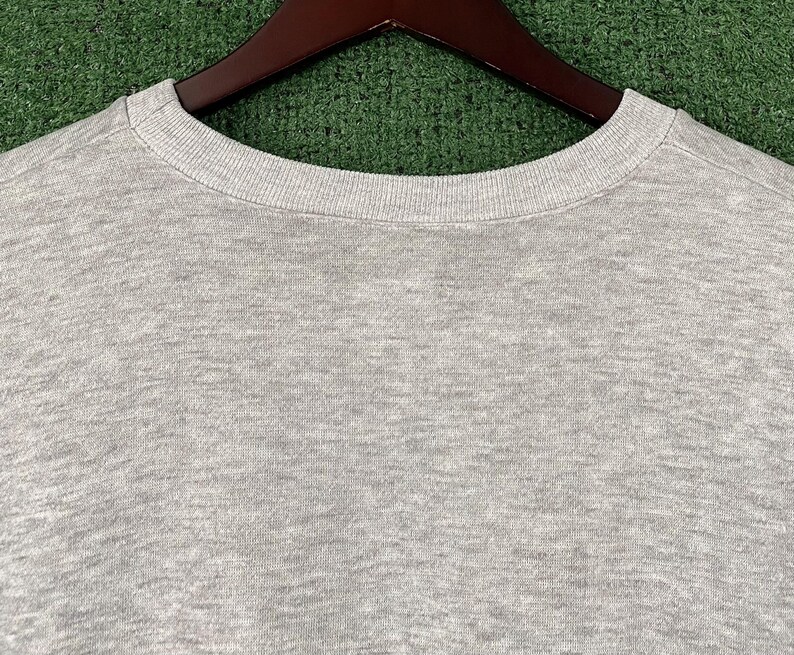 May include: A heather gray crew neck sweatshirt, displayed on a wooden hanger against a green background. The close-up shot highlights the texture of the fabric and the ribbed neckline. The sweatshirt is a casual apparel item.