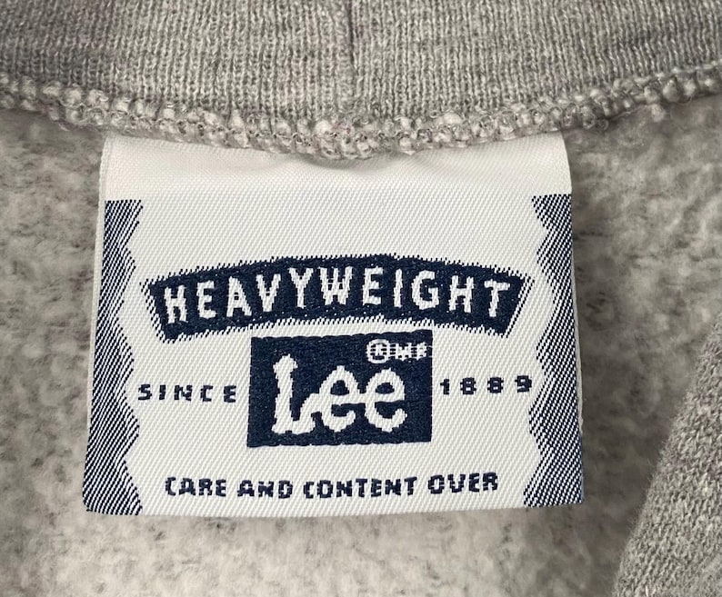 May include: Close-up of a grey sweatshirt collar with a white label. The label displays the text "HEAVYWEIGHT" and the Lee logo in navy blue. The label also includes the text "SINCE 1889" and "CARE AND CONTENT OVER".