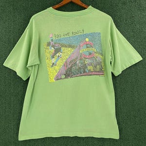 May include: A light green short-sleeve t-shirt featuring a graphic on the back. The design depicts a skateboarder and a steam locomotive with the text "RED HOT RAILS" and "TEAM INTERNATIONAL". The graphic uses yellow, purple, and blue colors.