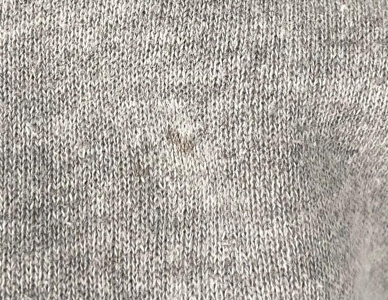May include: Close-up of a grey knitted fabric with a textured surface. The fabric appears to be a blend of light and dark grey yarns, creating a heathered effect. The knit pattern is visible, showing the individual stitches.