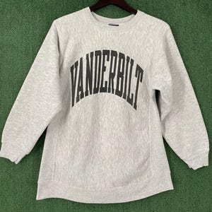 May include: A light gray sweatshirt with the word "VANDERBILT" printed in large, black, arched letters across the chest. The sweatshirt has a crew neck and long sleeves. The fabric appears to be a soft, comfortable material.