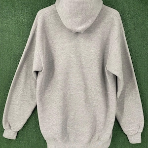 May include: A grey hoodie with a hood and long sleeves. The sweatshirt is hanging on a black hanger against a green background. The cuffs and bottom hem are ribbed.