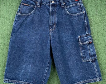 May include: Dark blue denim cargo shorts with multiple pockets and a button closure. The shorts have a relaxed fit and are hanging on a wooden hanger. The shorts have the brand name "FUBU" on the front pocket.
