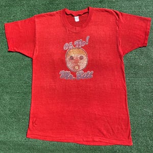 May include: A vintage red t-shirt with the words "Oh No! Mr. Bill" in a colourful design. The graphic includes a cartoon face with a dummy. The shirt has a crew neck and short sleeves.