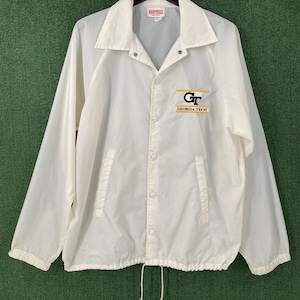 May include: A white nylon jacket with a collar and snap closures. The jacket features the Georgia Tech logo in gold and navy blue on the chest. The jacket has two front pockets and a drawstring at the bottom.