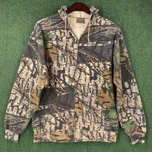 May include: A full-zip hooded sweatshirt with a camouflage pattern in shades of brown, gray, and green. The jacket has a drawstring hood, front pockets, and ribbed cuffs and hem. The sweatshirt is hanging on a wooden hanger.