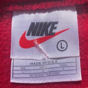Może przedstawiać: Zbliżenie białej metki z logo Nike, czerwonym znakiem Nike i nazwą marki w kolorze czarnym. Etykieta zawiera r&oacute;wnież rozmiar "L" i napis "MADE IN U.S.A.". Etykieta znajduje się na czerwonej tkaninie.