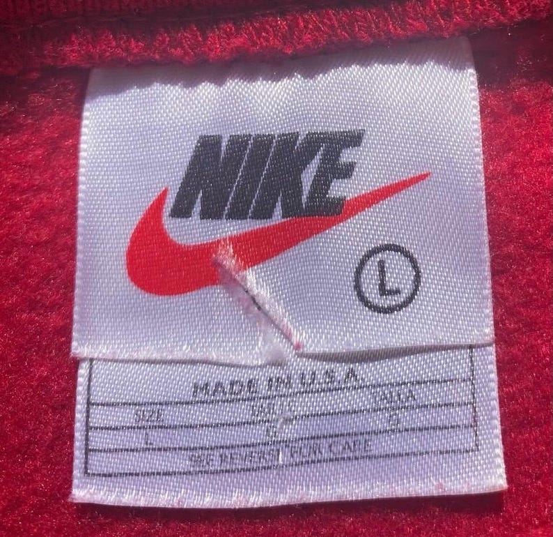 Może przedstawiać: Zbliżenie białej metki z logo Nike, czerwonym znakiem Nike i nazwą marki w kolorze czarnym. Etykieta zawiera r&oacute;wnież rozmiar "L" i napis "MADE IN U.S.A.". Etykieta znajduje się na czerwonej tkaninie.